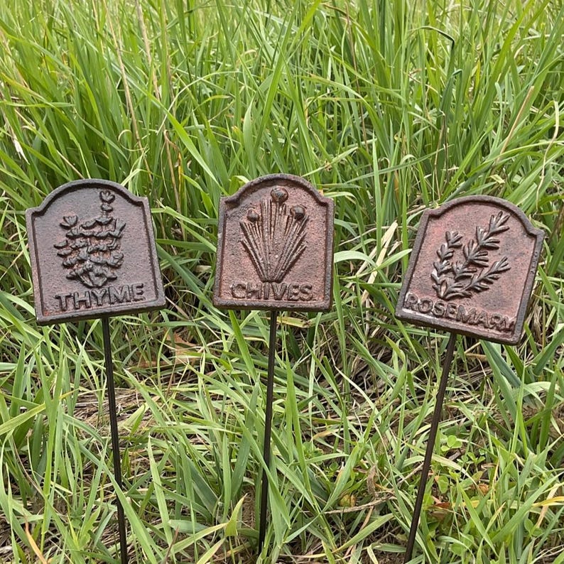 May include: Three weathered metal garden markers with the names "Thyme", "Chives", and "Rosemary" embossed on them. Each marker features a different embossed herb illustration. The markers are set in a grassy area.