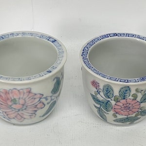 May include: Two white ceramic flower pots with blue and pink floral designs. The pots have a blue and white geometric border around the top.