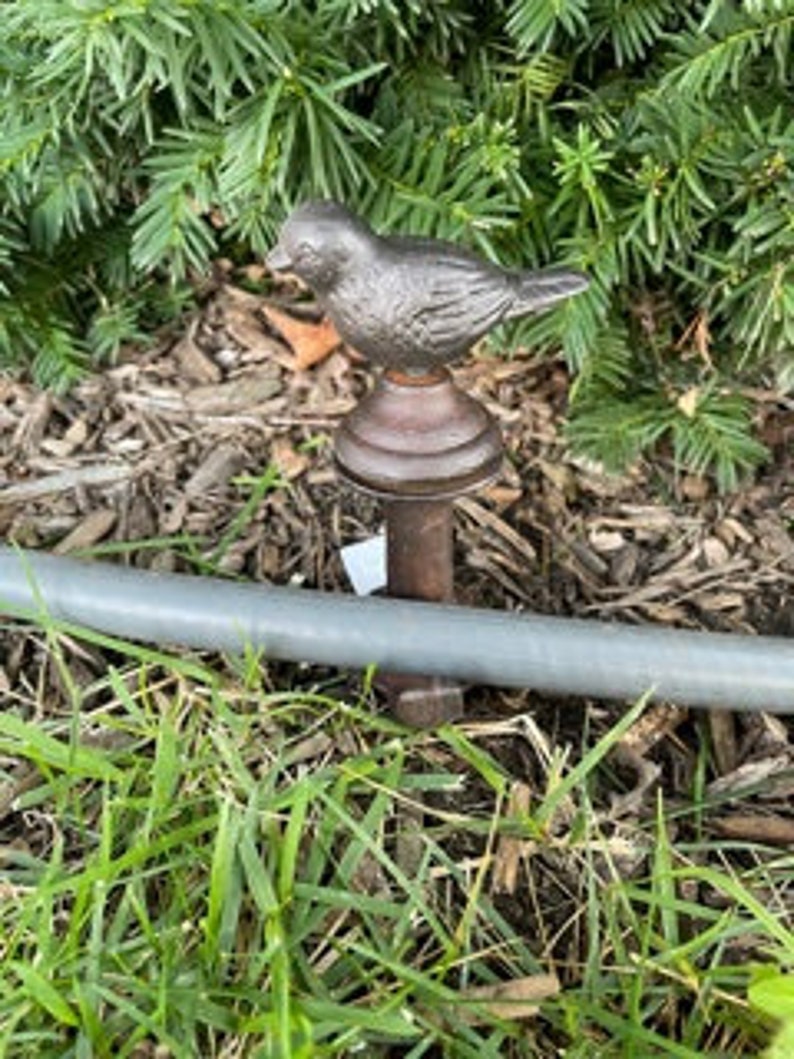May include: A bronze-colored bird finial atop a decorative rod. The bird is perched on a dark brown base, with a detailed feather design. The rod is a light gray color, and the background features green grass and brown mulch.