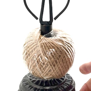 May include: A black metal twine holder with a decorative base and a scissor-shaped top. The holder supports a ball of natural-colored twine wrapped in clear plastic. The base has a detailed, ornate design.