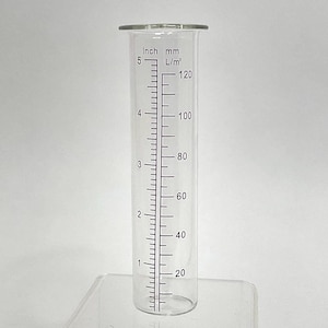 May include: A clear glass graduated cylinder with inch, mm, and L/m² markings. The cylinder is approximately 5 inches tall, with measurements up to 120 L/m². The cylinder has a wide, rounded top and is designed for measuring liquids.
