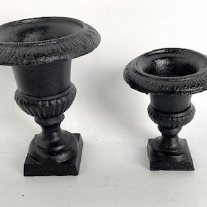 May include: Two black cast iron urns, one larger than the other, with a classic design. The urns have a round base and a wide, round opening.