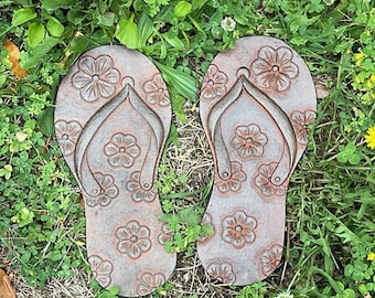 Op de afbeelding: Een paar verweerde stapstenen in de vorm van teenslippers. Elke steen heeft een bloemenpatroon en een teenband. De stapstenen hebben een gedempte bruine kleur en liggen in groen gras en klaver.