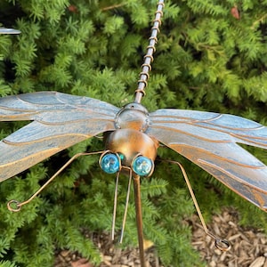 May include: A detailed metal dragonfly garden stake with blue glass eyes and intricate wing detailing. The dragonfly has a bronze and silver finish, with delicate antennae and legs. The wings are a light blue color. The garden stake is designed to be placed in a garden or outdoor space.