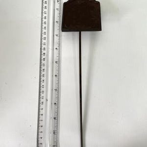 May include: A dark brown metal garden stake with a rectangular sign. The stake is approximately 30 cm long, with a ruler in the image for scale. The sign has a decorative top and is blank.