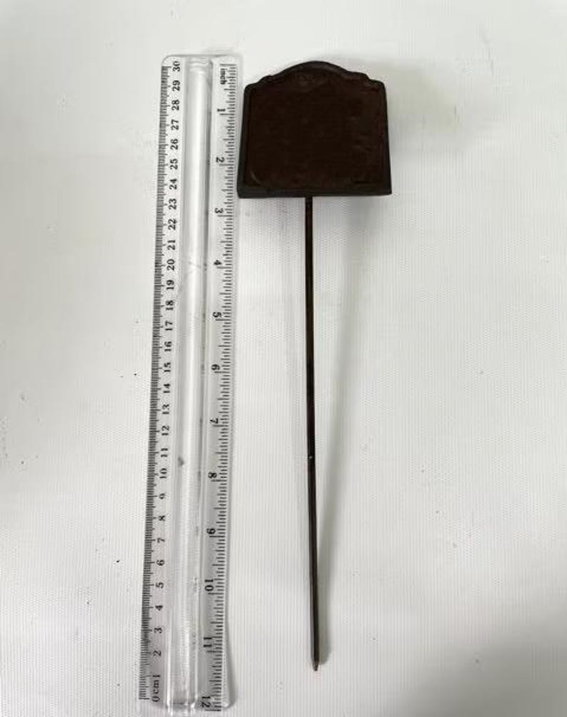 May include: A dark brown metal garden stake with a rectangular sign. The stake is approximately 30 cm long, with a ruler in the image for scale. The sign has a decorative top and is blank.