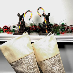 May include: Two black cast iron mouse figurines holding colorful striped candy canes, positioned behind two Christmas stockings with cream-colored cuffs and a silver-toned fabric body featuring a swirling pattern and snowflake motifs.
