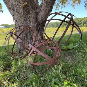 May include: Three large, spherical, metal garden art pieces with a rusty brown finish. The spheres are constructed from intersecting bands of metal, creating an open, geometric design. They rest on a bed of green grass near a tree.