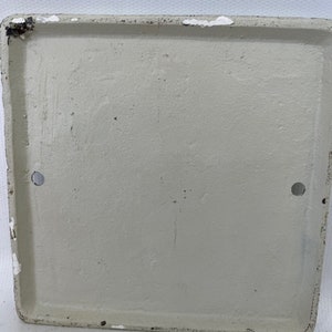 May include: A white, square, metal plaque with two small holes on the sides. The plaque is blank and has a slightly textured surface.