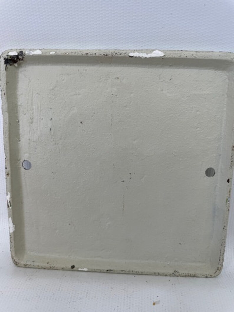 May include: A white, square, metal plaque with two small holes on the sides. The plaque is blank and has a slightly textured surface.