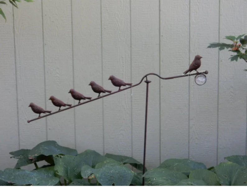 Pu&ograve; includere: Un paletto decorativo in metallo per giardino con sei piccole figurine di uccelli appollaiate su un'asta diagonale. Gli uccelli sono di colore marrone scuro e il paletto &egrave; posto su uno sfondo verde chiaro e beige.