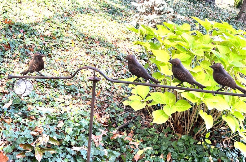 Pu&ograve; includere: Un paletto da giardino decorativo in metallo con quattro figurine di uccelli color bronzo appollaiate su un'asta curva. Il paletto include un piccolo specchio rotondo, il tutto su uno sfondo di verde.