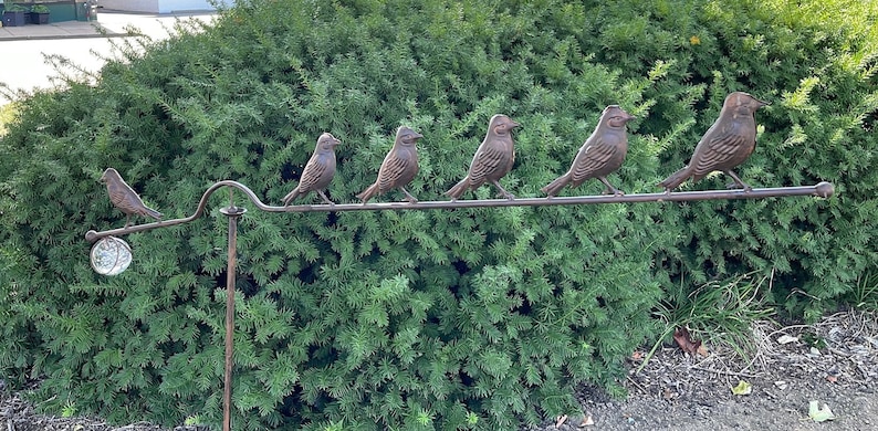 Pu&ograve; includere: Scultura decorativa in metallo raffigurante sette uccelli color bronzo appollaiati su un'asta curva. Gli uccelli sono disposti in fila. Lo sfondo &egrave; costituito da un fogliame verde, un elemento decorativo per il giardino.