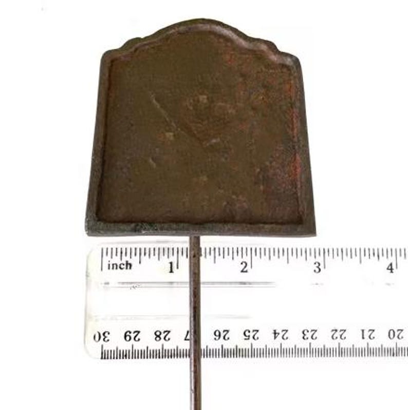 May include: A weathered, brown metal garden marker with a decorative top. The marker is approximately 7.6 cm tall and is attached to a thin metal rod. The background is white.