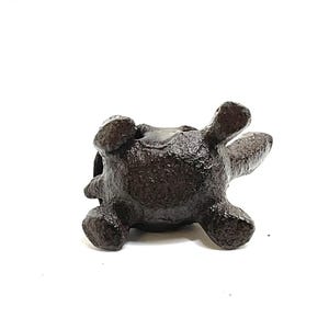 May include: A small, dark brown turtle figurine, seen from below, with its legs and head extended. The figurine has a textured surface, possibly metal. The turtle is set against a white background. A decorative item.