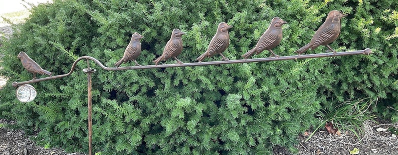 Pu&ograve; includere: Un paletto da giardino decorativo in metallo con sei sculture di uccelli color bronzo appollaiate su una barra orizzontale. Gli uccelli sono disposti in fila, con un globo di vetro all'estremit&agrave;. Il paletto &egrave; posto su uno sfondo di fogliame verde.
