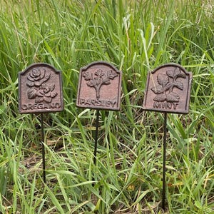 May include: Three weathered metal garden markers, each with a different herb name: "OREGANO", "PARSLEY", and "MINT". The markers feature embossed botanical illustrations of a rose, parsley, and mint leaves. They are set in tall green grass.