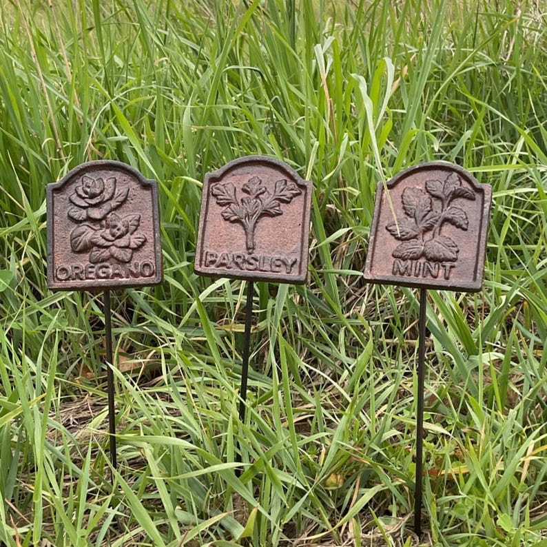 May include: Three weathered metal garden markers, each with a different herb name: "OREGANO", "PARSLEY", and "MINT". The markers feature embossed botanical illustrations of a rose, parsley, and mint leaves. They are set in tall green grass.