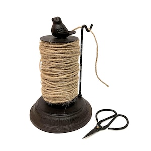 May include: A decorative twine dispenser with a small bird on top. The dispenser is made of dark metal and holds a spool of natural-colored twine. A pair of black scissors rests nearby, ready for use. The base is approximately 5 inches in diameter.