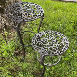May include: Two ornate, dark metal side tables with circular tops and intricate scrollwork designs. The tables have three legs and are set on a grassy lawn, showcasing a classic outdoor furniture style.