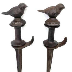 May include: Two cast iron bird garden stakes. Each stake features a small bird perched atop a decorative base, with a hook and pointed end for easy placement in the ground. The stakes are finished in a dark brown color.