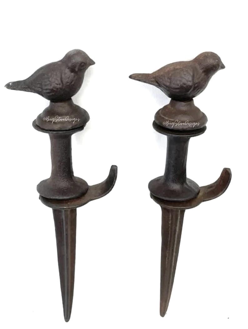 May include: Two cast iron bird garden stakes. Each stake features a small bird perched atop a decorative base, with a hook and pointed end for easy placement in the ground. The stakes are finished in a dark brown color.