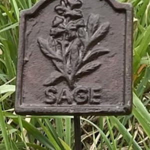 May include: A dark brown metal garden marker, rectangular in shape, with the word "SAGE" embossed. The marker displays a detailed relief of sage leaves and flowers. It is mounted on a thin metal stake, set against a backdrop of green grass.