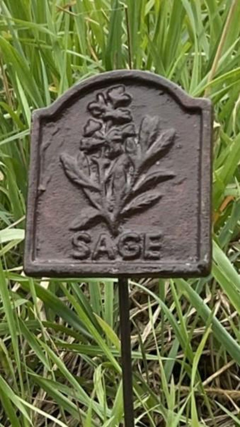 May include: A dark brown metal garden marker, rectangular in shape, with the word "SAGE" embossed. The marker displays a detailed relief of sage leaves and flowers. It is mounted on a thin metal stake, set against a backdrop of green grass.