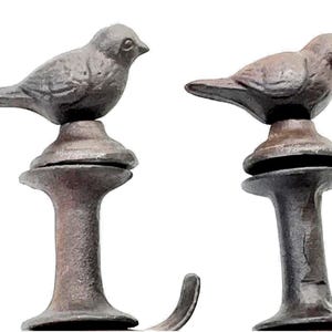 May include: Two cast iron bird-shaped decorative hooks. The birds are perched atop ornate, pedestal-style bases. The hooks are finished in a rustic, dark brown color, adding a vintage aesthetic. Ideal for hanging small items.
