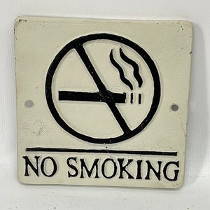 May include: White cast iron sign with black lettering that reads "NO SMOKING". A black and white circle with a diagonal line through it depicts a cigarette with smoke coming from it.