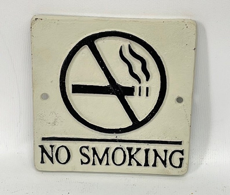 May include: White cast iron sign with black lettering that reads "NO SMOKING". A black and white circle with a diagonal line through it depicts a cigarette with smoke coming from it.