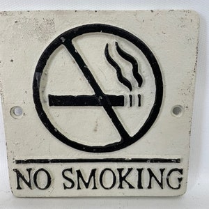 May include: White cast iron sign with black lettering that reads "NO SMOKING". A black circle with a diagonal line through it and a cigarette with smoke coming from it is above the text.