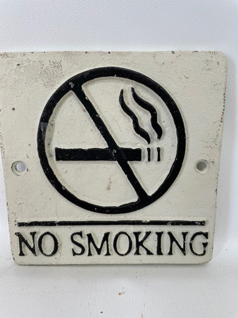 May include: White cast iron sign with black lettering that reads "NO SMOKING". A black circle with a diagonal line through it and a cigarette with smoke coming from it is above the text.