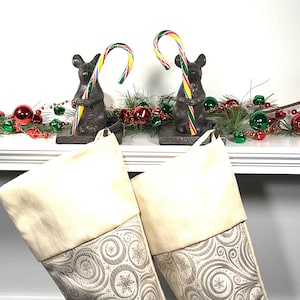 May include: Two Christmas stockings with cream-colored tops and patterned gray bottoms are displayed. Above, two dark gray mouse figurines hold colorful candy canes. A garland of red and green ornaments and greenery decorates the mantel.