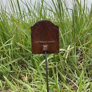 May include: A rectangular, weathered metal garden stake with a decorative top. The stake is a rusty brown colour and has the copyright symbol and the year 2025 printed on it. The stake is set in a field of tall green grass.