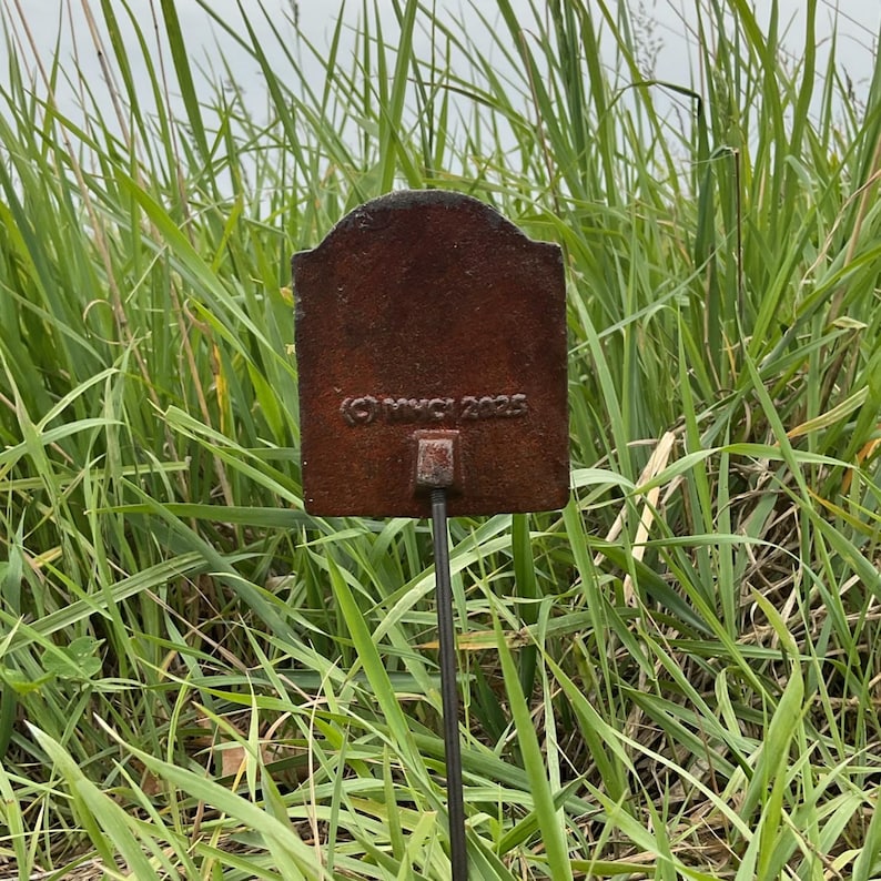 May include: A rectangular, weathered metal garden stake with a decorative top. The stake is a rusty brown colour and has the copyright symbol and the year 2025 printed on it. The stake is set in a field of tall green grass.