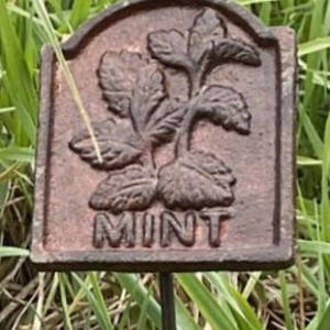 May include: A rectangular, bronze-coloured garden stake with a decorative arched top. The stake features a raised image of mint leaves and the word "MINT" in capital letters. The stake is set in a bed of green grass.