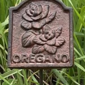 May include: A rectangular, weathered metal garden marker, featuring a floral design and the word "OREGANO". The dark, reddish-brown marker is mounted on a thin metal stake. Ideal for marking oregano plants in a garden, it is a decorative addition.