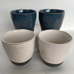 May include: Set of four ceramic mugs with a geometric pattern. Two mugs are teal blue with a black rim and two mugs are white with a black rim.