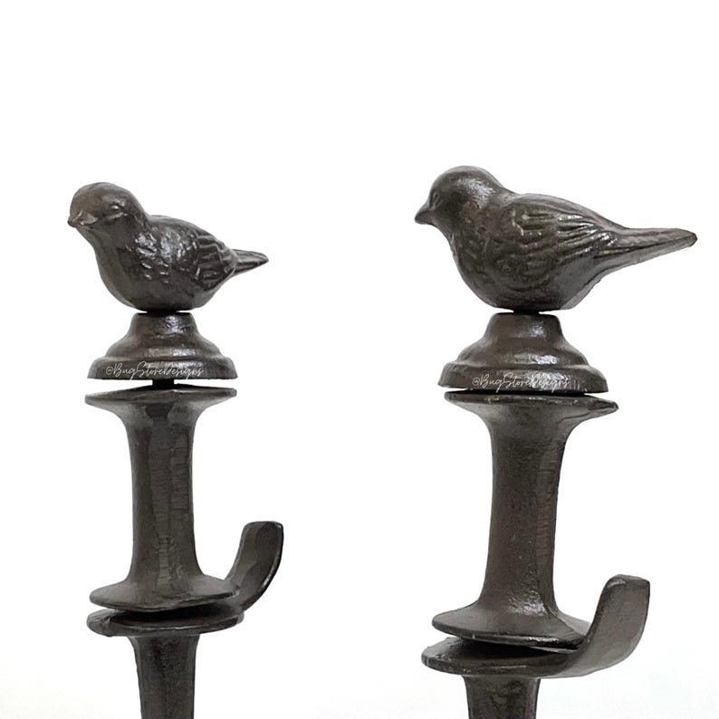May include: Two dark brown metal hooks with a bird perched on top. The hooks have a decorative design and are suitable for hanging items. The birds are detailed and add a touch of nature to the design.