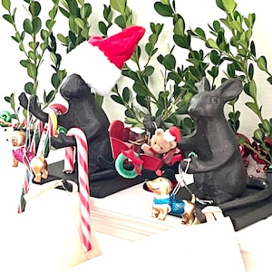 May include: Two dark gray mouse figurines, one wearing a red and white Santa hat, pulling a red sleigh containing a small teddy bear. The mice are decorated with red and white candy canes and a small tan dog figurine wearing a blue glitter sweater.