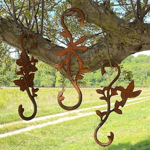 May include: Three decorative metal garden hooks with an aged, rusty finish. Each hook features a different nature-inspired design: butterflies, a dragonfly, and a hummingbird with leaves. The hooks are suspended from a tree branch against a backdrop of green grass and trees.