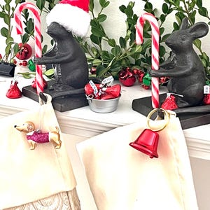 May include: Two black cast iron mouse figurines holding red and white striped candy canes. One mouse wears a red and white Santa hat. The candy canes support two cream-colored stockings, one featuring a dachshund ornament and the other a red bell.