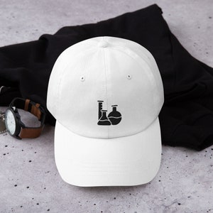 Peut inclure: Casquette de baseball blanche avec un motif noir de verrerie de laboratoire, comprenant un bécher, une fiole et une éprouvette graduée. Le chapeau est en matière souple et possède une visière incurvée. Style classique, pour un usage décontracté.