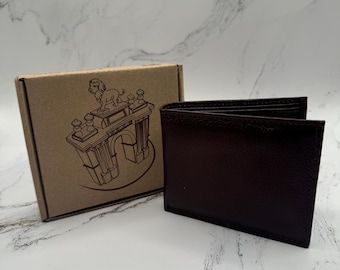 May include: A dark brown leather bifold wallet positioned next to a square cardboard gift box. The box features a black line drawing of a lion standing atop an arched monument. The wallet has a textured surface and is shown in a partially open state.