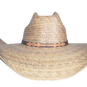 K&ouml;nnte beinhalten: Ein breitkrempiger Strohhut im Cowboy-Stil mit einem braunen Lederband und dekorativen Metallverzierungen. Der Hut hat eine hellbraune Farbe und eine leicht spitze Krone.