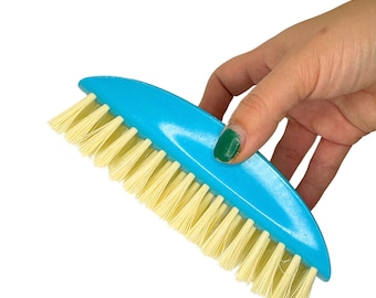 May include: A blue plastic brush with a curved handle and yellow bristles. The brush is being held in a hand.