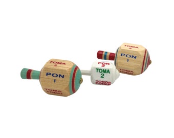 May include: Three wooden spinning tops with different colored stripes and text in Spanish. The tops have a white, green, and red color scheme. The text on the tops reads "Toma 2", "Pon 1", and "Toma Todo".