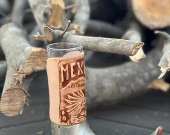 May include: A tall shot glass encased in a silver-toned, pointed-toe boot-shaped holder. The upper portion features a tan leather sleeve with an embossed design of a sun, mountains, and the text MEX, showcasing detailed craftsmanship.