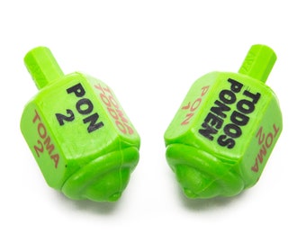 May include: Two green plastic dreidels with black and red text. The text includes the words "PON", "TOMA", and "TODOS PONEN". The number "2" is also visible on the dreidels. The dreidels are set against a white background.